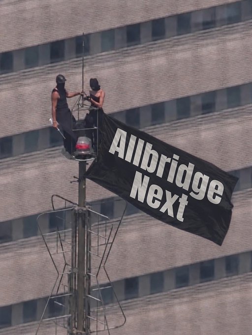 Two people on top of a communications tower unfurling a black banner that reads 'Allbridge Next' against the backdrop of a high-rise building.