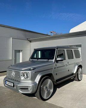 A silver Mercedes-Benz G-Class SUV parked in front of a building with gray siding and glass doors, bearing the license plate 'ME G 780E'.