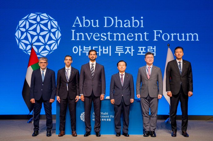 Five men in suits pose for a group photo at the 'Abu Dhabi Investment Forum' with a UAE flag and a Japanese flag visible.