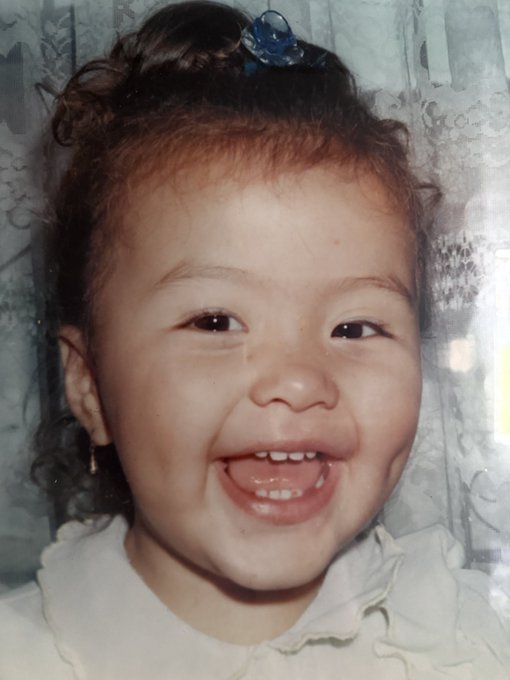A vintage photograph of a smiling young child with curly brown hair and a blue hair accessory, wearing a white ruffled top.