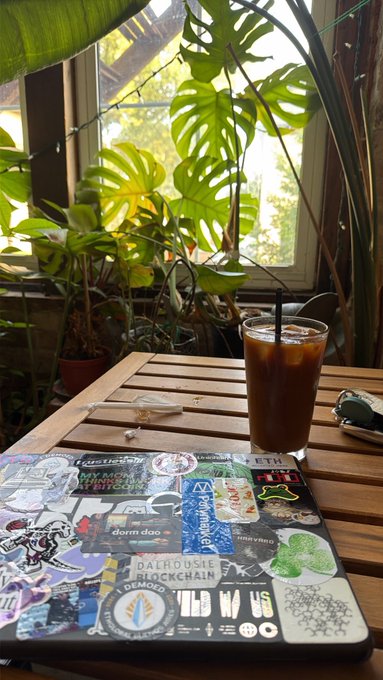 A laptop covered in tech and crypto stickers sits on a wooden table next to a glass of iced coffee, with lush green plants visible through a window in the background.