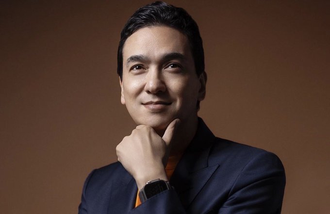 A professional portrait of an Asian man in a navy blue suit with an orange tie, resting his chin on his hand and wearing a smartwatch against a warm brown background.