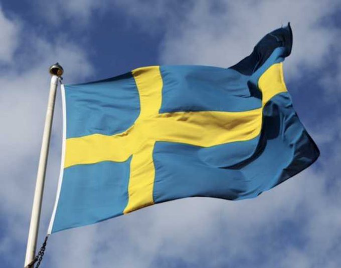 The Swedish national flag, featuring a yellow Nordic cross on a blue field, waving against a partly cloudy sky.