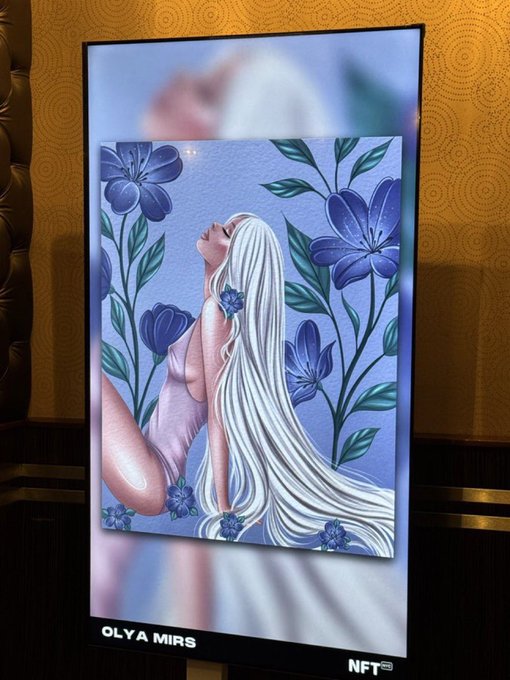 A digital display screen shows an NFT artwork by OLVA MIRS, depicting a pale figure with flowing white hair surrounded by blue flowers and a crescent moon.