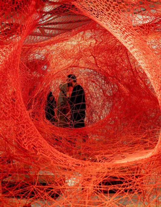 A person viewed through the center of an intricate, web-like red tunnel art installation, creating a hypnotic circular perspective.
