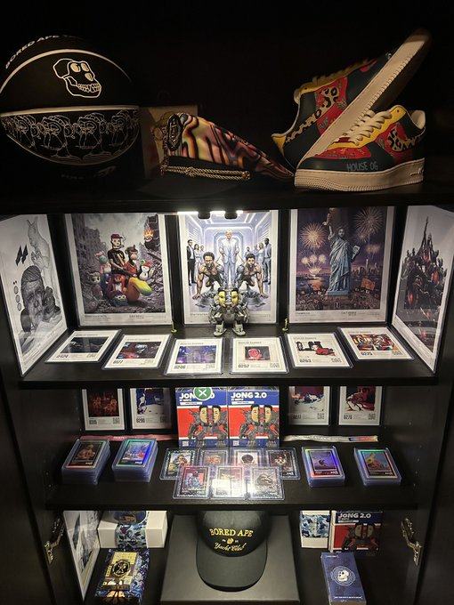 A display case showcasing a "Bored Ape" themed collection featuring Beeple TCG cards, numbered posters, jerseys, and branded sneakers including "Mike O" sneakers, with "JONG 2.0" art cards visible—reflecting the post's enthusiasm for Beeple TCG collecting and NFT-inspired memorabilia.
