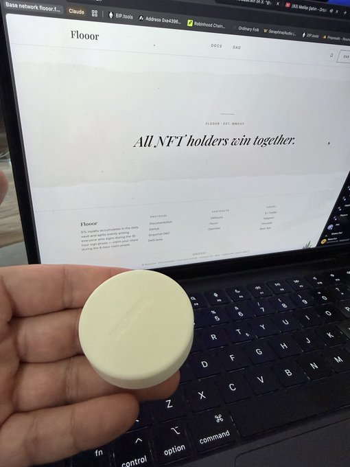 Hand holding a pale yellow circular puck-like object with an embossed logo in front of a laptop displaying a 'FLOOR' NFT website with text 'All NFT holders win together.'