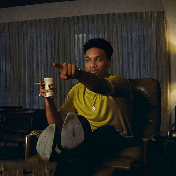 A young man in a yellow shirt sitting in a chair holding a coffee mug in one hand and pointing forward with the other, in a dimly lit room.