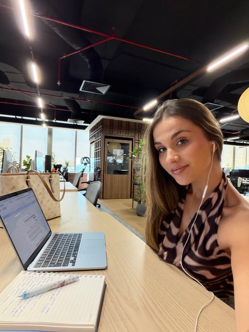 A 22-year-old content creator and memecoin trader from Spain, based in Thailand, takes a selfie while working at her laptop and notebook in a modern co-working space. The setting—featuring an open laptop with trading content, a handwritten notebook, and earbuds—reflects her lifestyle of blending digital entrepreneurship with the crypto trading space she mentions wanting to grow within.