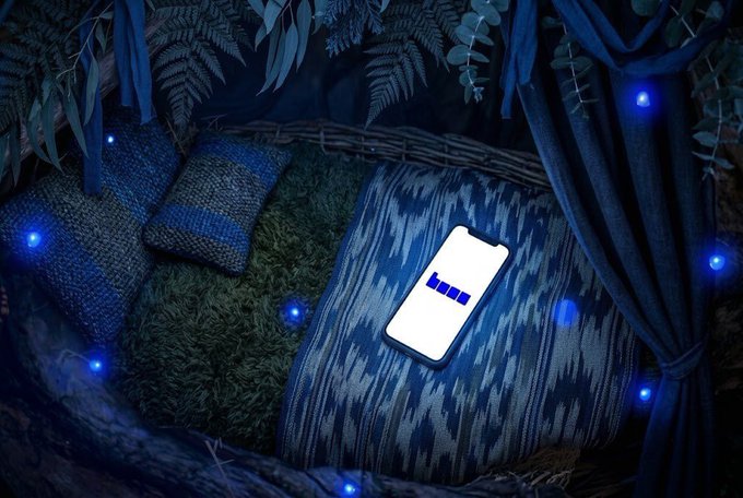 A smartphone displaying the Base logo rests on a cushion inside a cozy, dimly lit nook surrounded by ferns, woven baskets, and small blue lights, evoking a sense of quiet focus and late-night building. The intimate, blue-toned setting mirrors the post's message about the power of "quiet builders" who grow steadily outside the spotlight, channeling the same understated, focused energy into contributing to @base and @jessepollak's vision.