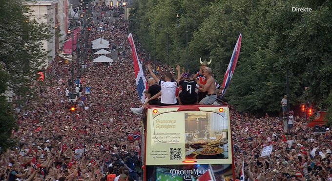 An aerial view of a large crowd gathered in a city street, with people on top of a double-decker bus displaying 'Buoranza Eveliquiet' and 'PrideMap Pub' signage, celebrating with British flags.