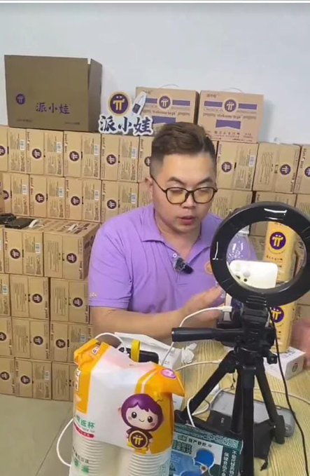 A man in a purple polo shirt and glasses is filming a live stream, with stacked product boxes labeled "派小娃" visible in the background and a ring light on a tripod in front of him. He appears to be promoting or demonstrating paper cups from a brand, reinforcing the post's message about prioritizing practical usefulness in products.