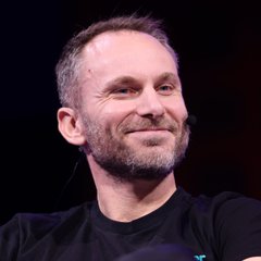 A close-up portrait of a smiling man in a dark shirt against a black background.