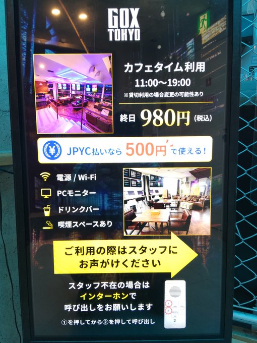 Photo of an illuminated GOX TOKYO café advertisement in Japanese, listing café time hours 11:00-19:00, a 980 yen fee (500 yen with JPYC), amenities, and intercom instructions.