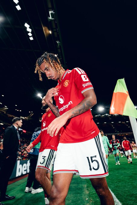 A Manchester United soccer player wearing jersey number 15 in a red kit with Snapdragon and summit sponsorships, shown on the field near another teammate wearing number 3 with an assistant and fans in the background.