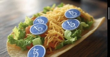 A taco filled with lettuce, cheese, and tomatoes is topped with blue coins bearing the Tezos "tz" symbol, playfully linking the food to a Tezos-themed art post for "Tezos Tuesday."
