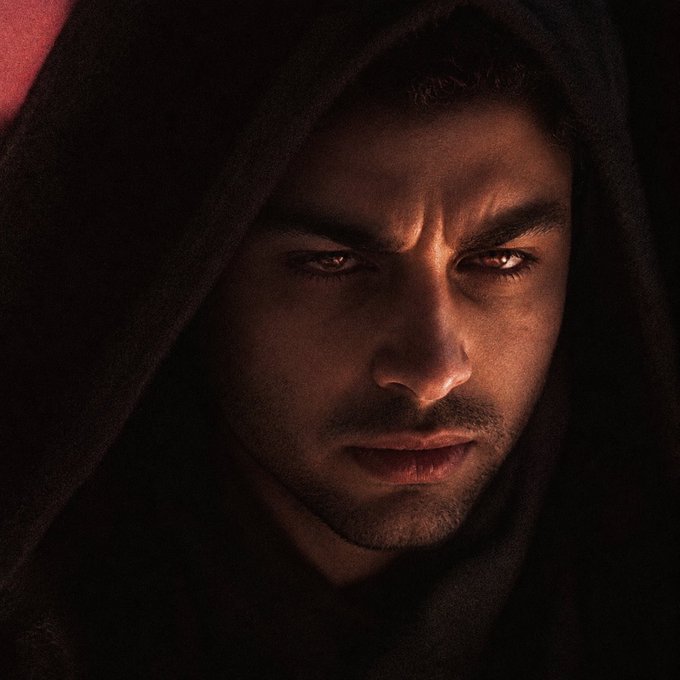 A dramatic, dimly lit portrait of a man with intense, glowing red eyes and facial scarring, wearing a dark hood.