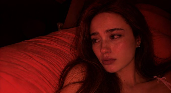 Close-up portrait of a woman with long brown hair lying on a bed, illuminated by red lighting, with a sorrowful expression.