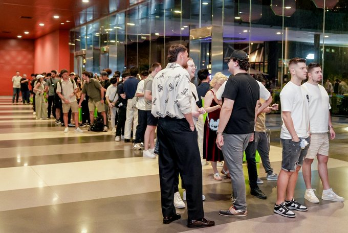 A crowd of people stands in a line within a modern building featuring large glass windows and warm interior lighting.