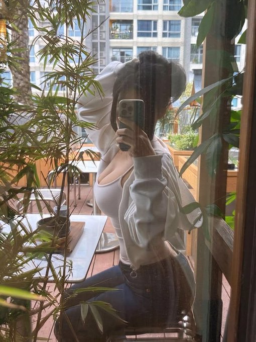 A mirror selfie of a woman wearing a white crop top, open white shirt, and jeans, holding a phone on a balcony surrounded by bamboo plants.