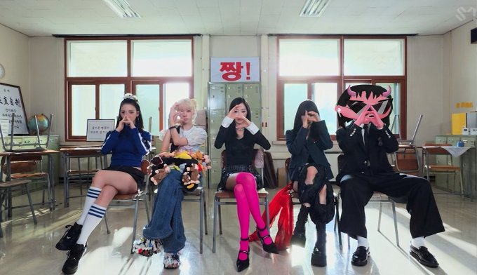 Five members of a K-pop group posing in a classroom setting with Korean text '짱!' (best/awesome!) displayed on screen, featuring one member with a censored face.