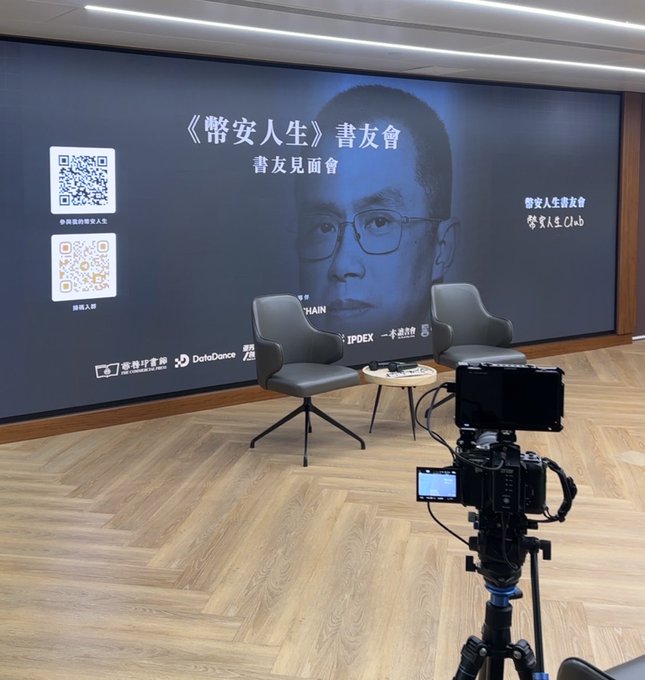The image shows a stage setup for a "《幣安人生》書友會" (Binance Life Book Club) event in Hong Kong, featuring a large backdrop with a portrait, two chairs around a coffee table, and a professional camera filming the scene—consistent with the user's arrival to support CZ at a Binance-related book event.