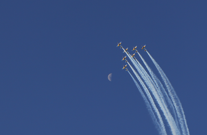 A formation of jets flies through a clear blue sky, leaving white smoke trails, with a crescent moon visible in the background.