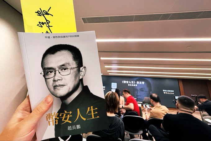 A hand holds up a Chinese-language memoir titled "幣安人生" (Binance Life) by 趙長鵬 (Changpeng Zhao), featuring his black-and-white portrait on the cover, at what appears to be a book launch event with attendees seated in the background. The image directly reflects the post about Binance Life being CZ's memoir, showcasing the physical book at the launch.