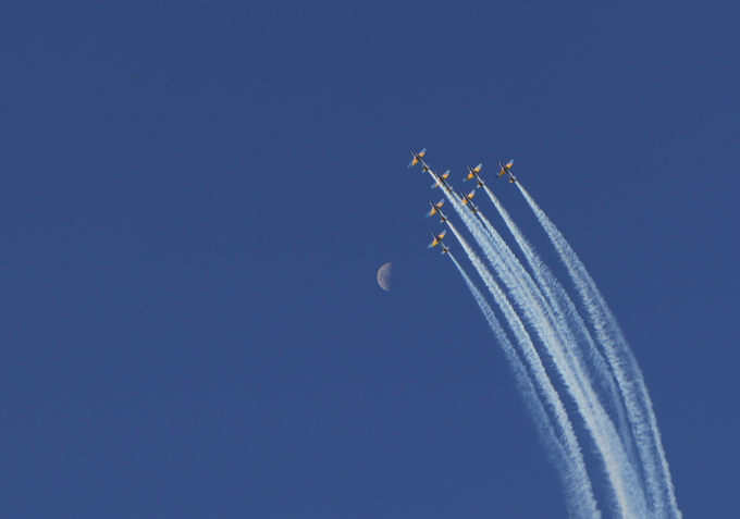 Six jets fly in formation leaving white contrails against a deep blue sky, with a crescent moon visible in the background.