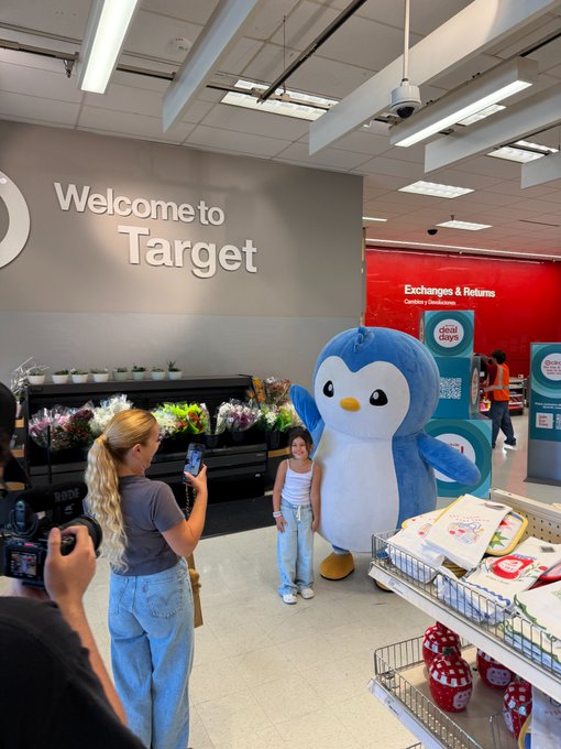 A woman poses with a large penguin mascot inside a Target store beneath a "Welcome to Target" sign, while another woman takes her photo. The scene connects to the caption "Your entire bloodline will Huddle," referencing Target's iconic mascot bullseye circle and the brand's playful, family-friendly image.