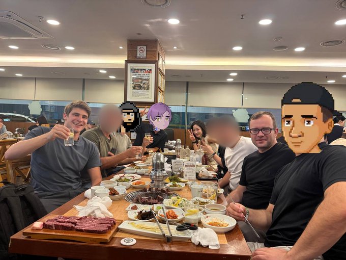 A group of friends and colleagues gather around a table filled with Korean BBQ dishes, raising glasses in a toast during a casual dinner outing together.
