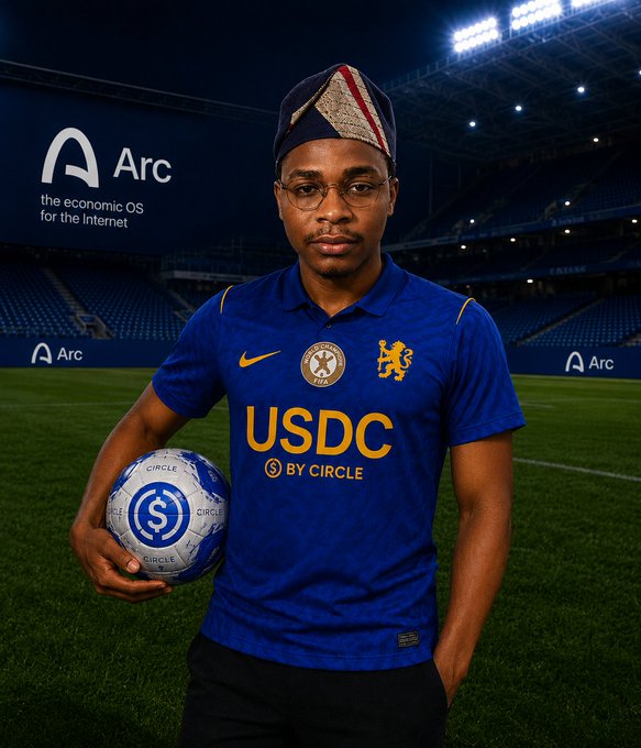 A young man wearing glasses and a traditional military cap poses on a football pitch wearing a blue jersey with "USDC BY CIRCLE" sponsorship, holding a branded football, with "Arc" advertising boards in the background.