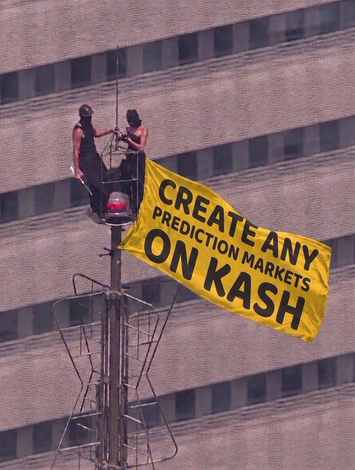 Two people stand on a platform atop a communications tower against a brick building backdrop, unfurling a yellow banner reading 'CREATE ANY PREDICTION MARKETS ON KASH'.
