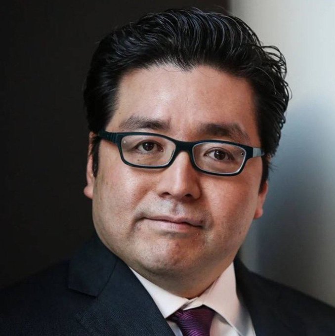 A professional headshot shows a man wearing dark rectangular frames, a dark suit, a white shirt, and a dark red tie against a gray background.
