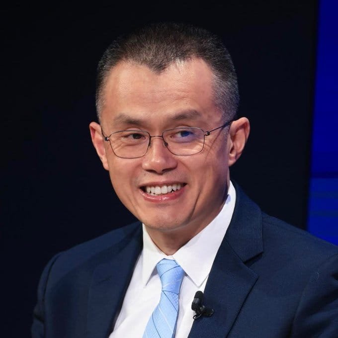 A headshot portrait of a smiling middle-aged man wearing glasses, a dark suit, and a light blue tie against a dark background.