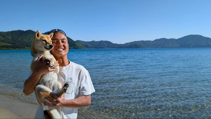 A young man in a white t-shirt smiles while holding a Shiba Inu dog on a beach, with calm blue water and forested mountains in the background.