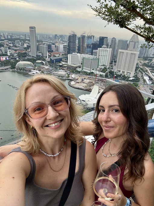 Two women smile for a selfie while holding wine glasses at a rooftop bar overlooking the Singapore skyline, including the Marina Bay area, as the poster prepares to attend Token2049 and connect with marketers working in crypto treasury operations.