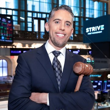 The image features a man dressed in a suit, standing confidently with his arms crossed. He is smiling and posing for a photo, positioned between two display screens showcasing a large amount of data from Strive's Bitcoin accumulation hit during a single day on January 11th, 2016 - approximately 50% of the previous week's total BTC haul. In the background, there are more financial indicators displayed on the screens behind him.