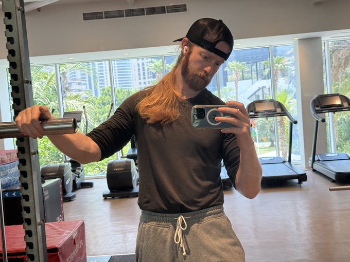 A long-haired man with a beard wearing a black cap and long-sleeve shirt takes a mirror selfie with his phone while holding a barbell in a gym with treadmills in the background.