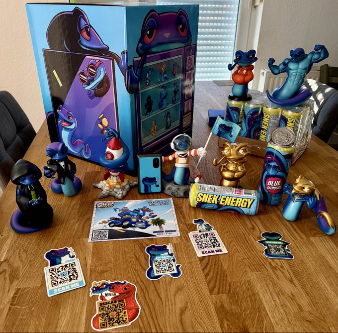 A wooden table displays a collection of blue snake-themed merchandise, including figurines, "SNEK ENERGY" cans, stickers with QR codes saying "SCAM ME," and an iPhone case, with a matching box in the background.