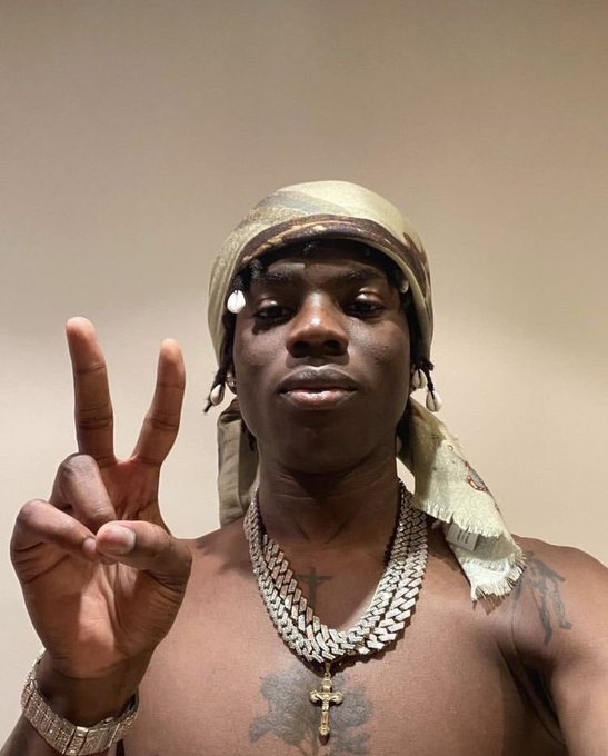 A shirtless man wearing a patterned headwrap and layered diamond chains with a cross pendant holds up a peace sign.