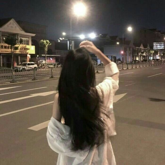 A woman with long black hair, wearing a white off-shoulder top, stands on a city street at night with her back to the camera, raising her arm as if hailing a taxi.