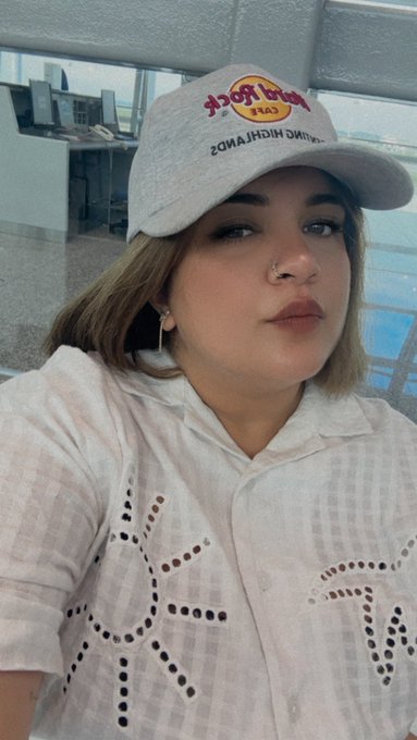 A selfie of a young woman wearing a white checkered shirt and a light gray Hard Rock Cafe cap, with a nose piercing and blonde shoulder-length hair.