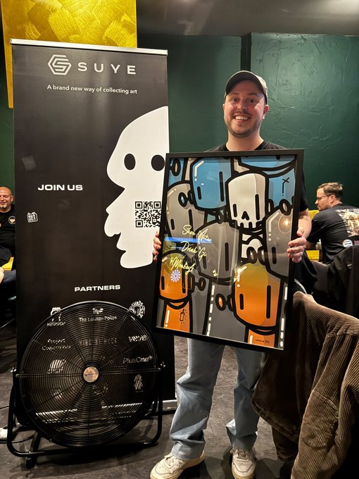 A man wearing a cap smiles while holding a framed colorful abstract artwork in front of a 'SUVE' banner advertising 'A brand new way of collecting art' alongside a large fan.