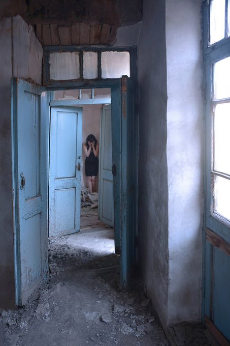 A figure stands in a doorway of an abandoned building with peeling teal paint, hands covering their face, framed by weathered doors and debris-strewn floors.