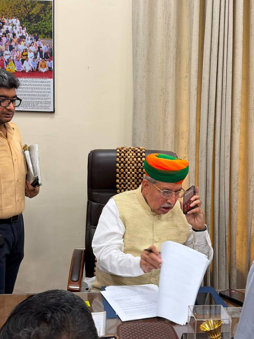 An older Indian man wearing a traditional colorful Rajasthani turban and beige vest, seated at a desk while talking on a mobile phone and reviewing documents, with an assistant standing nearby.