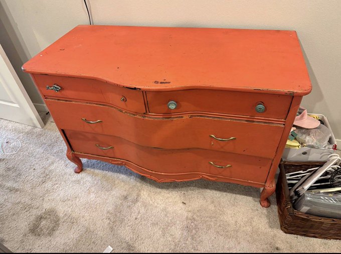 A distressed coral-orange vintage dresser with curved drawers and cabriole legs sits on a beige carpet next to a wicker basket.