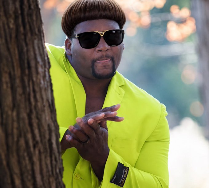 A man with a distinctive bowl haircut wearing dark sunglasses and a bright neon green blazer poses near a tree, with a 'SKINNY' visible on his sleeve tag.