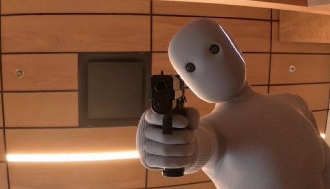 A small white robot with button eyes points a handgun directly at the camera in an indoor setting.