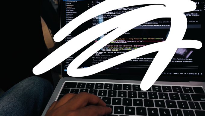 A person's hand types on a laptop keyboard displaying code, with a white scribble overlay obscuring part of the screen.
